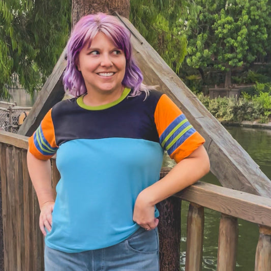 Person with purple hair standing on a wooden bridge with greenery in the background wearing a Goofy Disney themed shirt