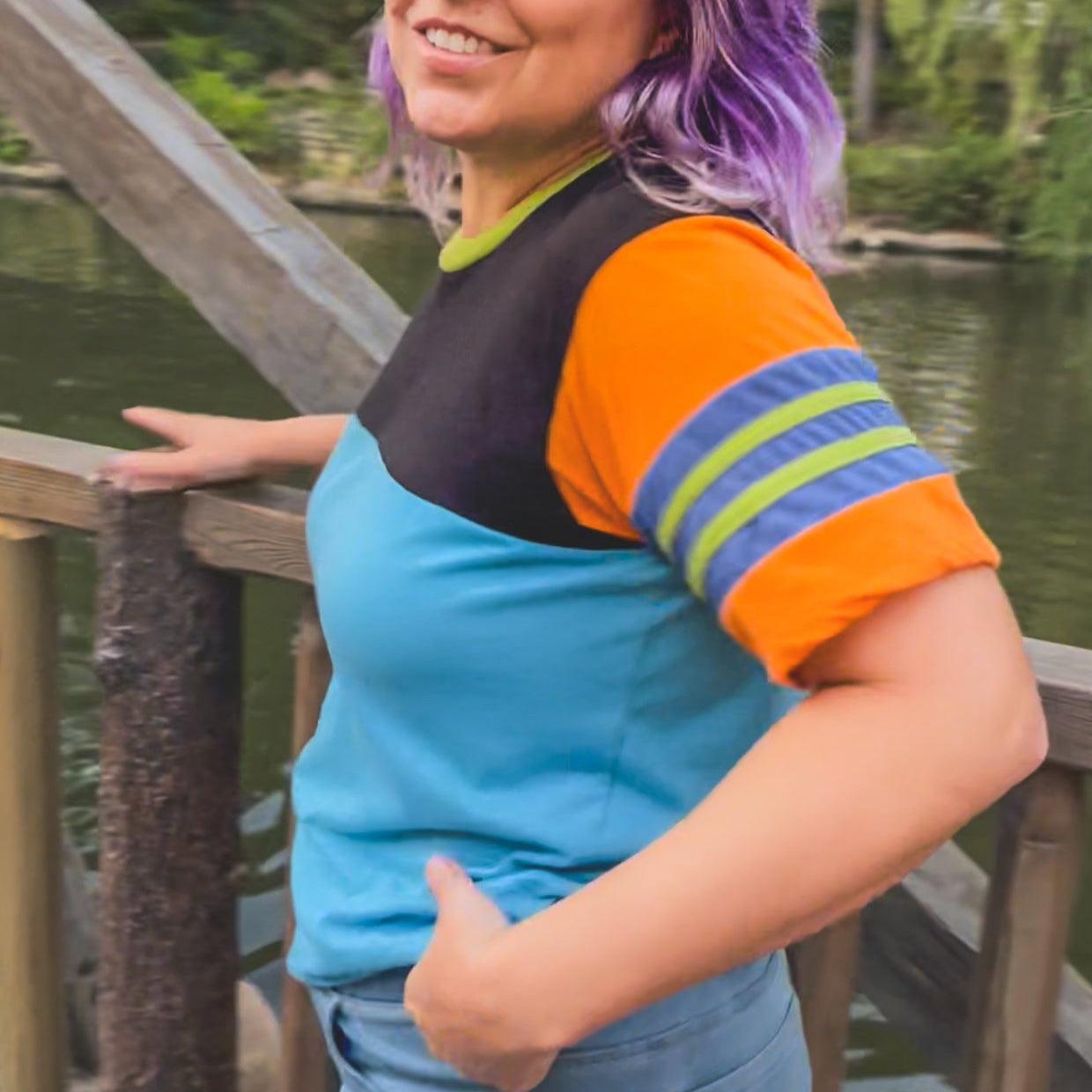 Person with colorful hair standing by a wooden railing with a scenic background at Disneyland  wearing a black, blue, orange, and green Goofy themed shirt 
