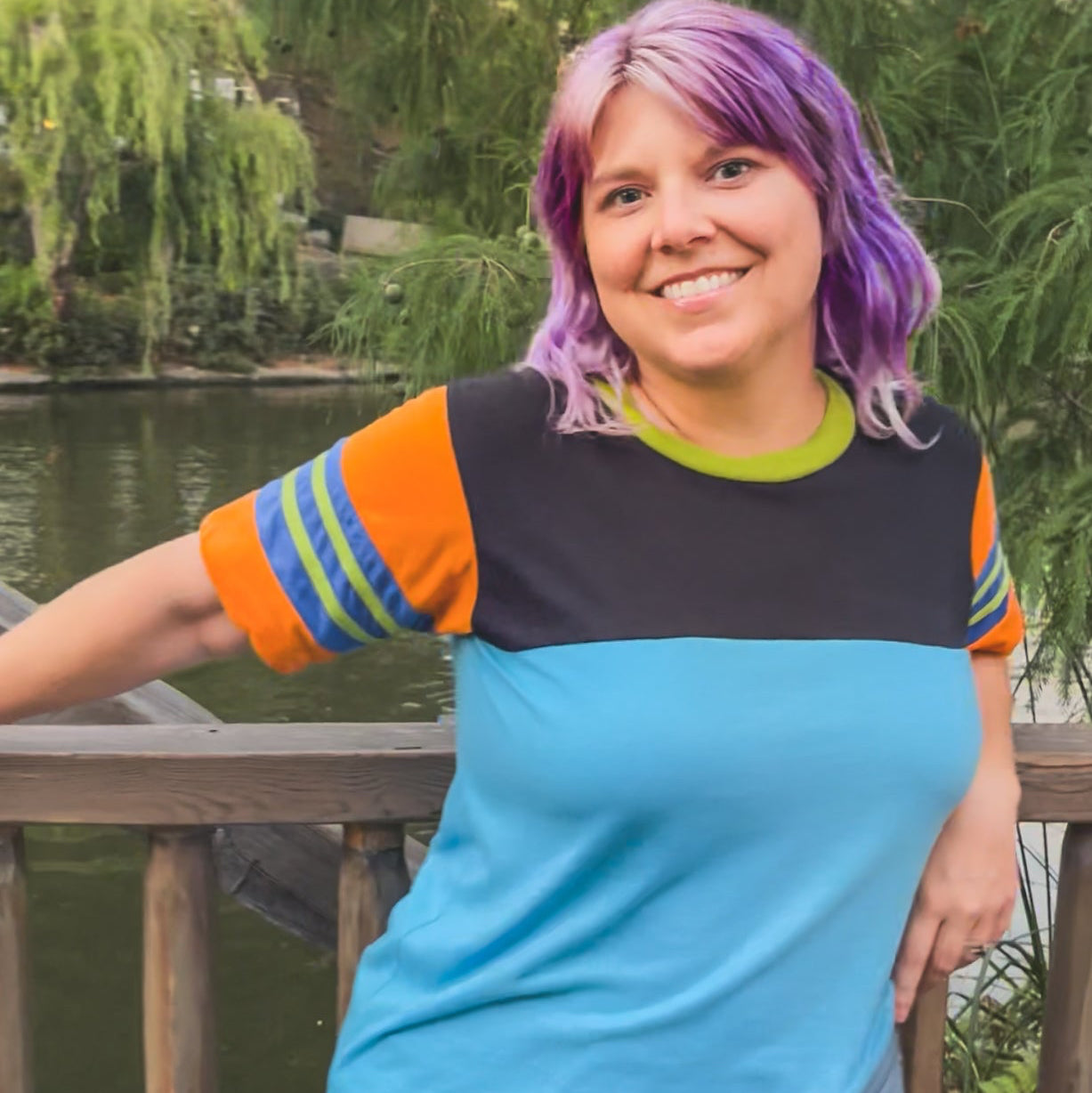 Person with colorful hair standing by a wooden railing with a scenic background at Disneyland  wearing a black, blue, orange, and green Goofy themed shirt 