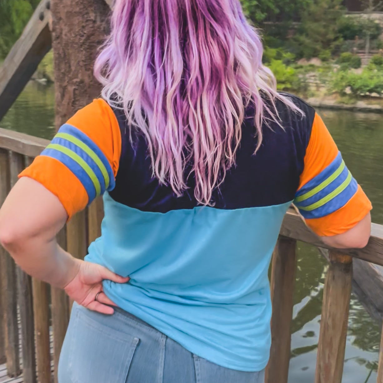 Person with colorful hair standing by a wooden railing with a scenic background at Disneyland  wearing a black, blue, orange, and green Goofy themed shirt 